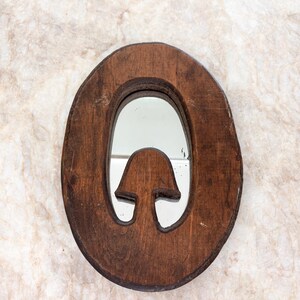 May include: An oval-shaped wall mirror with a dark brown wooden frame. The frame features a cutout design in the lower center that resembles a mushroom shape, which is integrated into the mirror's reflective surface area.