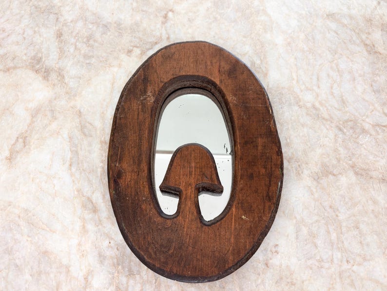 May include: An oval-shaped wall mirror with a dark brown wooden frame. The frame features a cutout design in the lower center that resembles a mushroom shape, which is integrated into the mirror's reflective surface area.