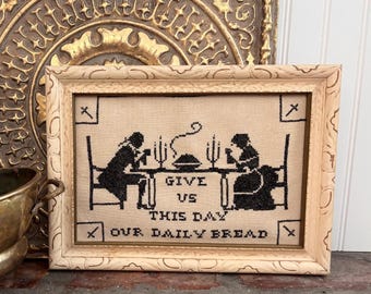 Op de afbeelding: Een ingelijste kruissteek met zwart garen op een beige stoffen achtergrond. Het ontwerp toont twee figuren aan een tafel met een dampend gerecht en kaarsen, vergezeld van de tekst: GIVE US THIS DAY OUR DAILY BREAD.