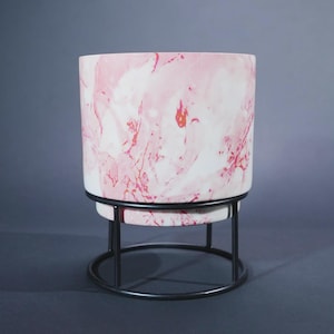May include: A cylindrical planter featuring a white and pink marbled pattern with swirling colours. The planter is supported by a black metal stand consisting of a circular base and three vertical rods that hold the pot in an elevated position.