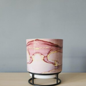 May include: A cylindrical planter featuring a marbled pattern in shades of dusty pink, deep burgundy, and gold accents. The planter is supported by a black metal wire stand with a circular base and four vertical supports holding the pot.