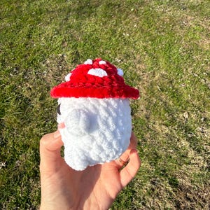 K&ouml;nnte beinhalten: Ein handgefertigtes H&auml;kelpilz-Spielzeug mit rotem Hut und wei&szlig;en Punkten. Der Pilz hat einen wei&szlig;en K&ouml;rper und ein kleines Gesicht. Das Spielzeug wird in einer Hand vor einem Hintergrund aus gr&uuml;nem Gras gehalten.