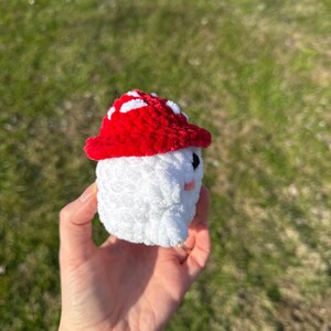 K&ouml;nnte beinhalten: Ein handgefertigtes geh&auml;keltes Pilzspielzeug mit rotem Hut mit wei&szlig;en Punkten und wei&szlig;em K&ouml;rper. Der Pilz hat kleine schwarze Augen und rosa Wangen. Das Spielzeug wird in einer Hand vor einem verschwommenen gr&uuml;nen Hintergrund gehalten.
