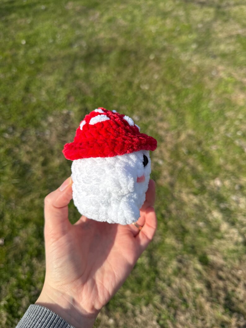 K&ouml;nnte beinhalten: Ein handgefertigtes geh&auml;keltes Pilzspielzeug mit rotem Hut mit wei&szlig;en Punkten und wei&szlig;em K&ouml;rper. Der Pilz hat kleine schwarze Augen und rosa Wangen. Das Spielzeug wird in einer Hand vor einem verschwommenen gr&uuml;nen Hintergrund gehalten.