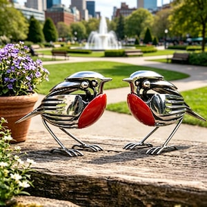 May include: Two polished metal bird sculptures with red breasts and detailed feather designs. The birds are perched on thin metal legs, facing each other. The background features a park with a fountain and buildings.