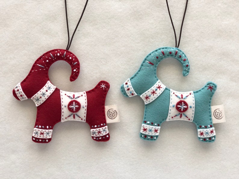 May include: Two felt Christmas ornaments shaped like goats. One is red with white accents and the other is blue with white accents. Both have a white star on their back.