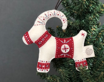 May include: A handmade Christmas goat ornament. The white felt goat has red and silver embroidered details. The ornament is suspended by a brown string and is set against a green Christmas tree. The goat is decorated with red and silver embroidery.