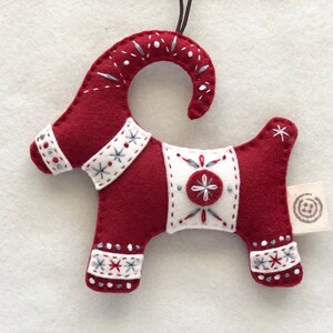 May include: Red felt Christmas ornament shaped like a goat with white and gray embroidered details. The ornament has a loop for hanging and a small white tag with a gray circle design.