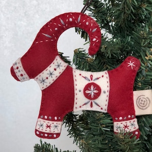 May include: Red felt Christmas ornament shaped like a goat with white and gray embroidered details. The ornament has a loop for hanging and a small tag with a circular design.