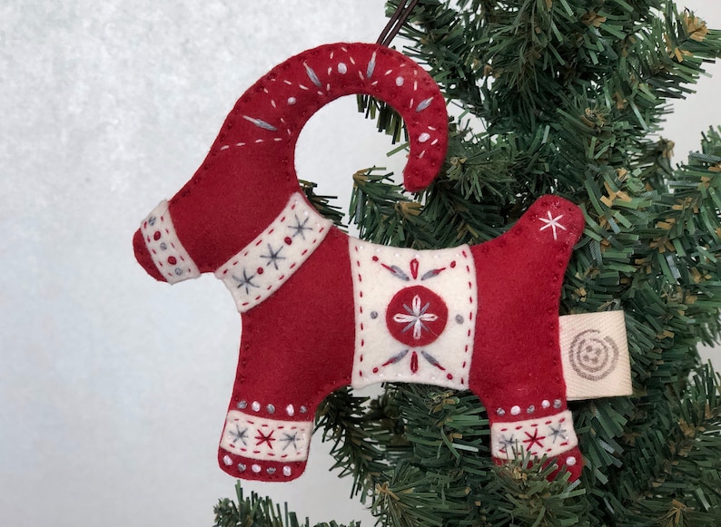 May include: Red felt Christmas ornament shaped like a goat with white and gray embroidered details. The ornament has a loop for hanging and a small tag with a circular design.