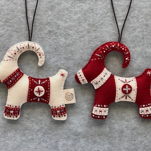 May include: Two felt Christmas ornaments shaped like goats. One goat is white with red accents and the other is red with white accents. Both goats have embroidered details and a loop for hanging.