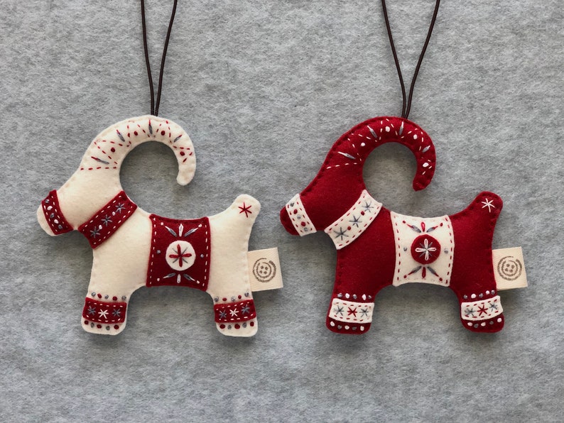 May include: Two felt Christmas ornaments shaped like goats. One goat is white with red accents and the other is red with white accents. Both goats have embroidered details and a loop for hanging.