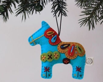 May include: A blue felt Christmas ornament shaped like a horse with colorful felt accents and embroidery. The ornament is hanging from a black string.