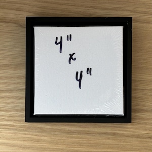 May include: Square white canvas in a black frame. The canvas is labeled with the measurements "4" x 4"" in black marker. The canvas is wrapped in clear plastic.