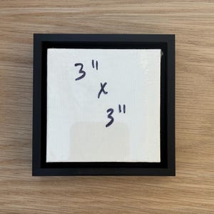 Puede incluir: Un pequeño lienzo cuadrado con el texto escrito a mano "3" x "3" en rotulador negro, enmarcado en un marco negro. El lienzo es blanco y parece estar estirado. El marco es de diseño sencillo y moderno. El lienzo mide 7,62 cm x 7,62 cm.