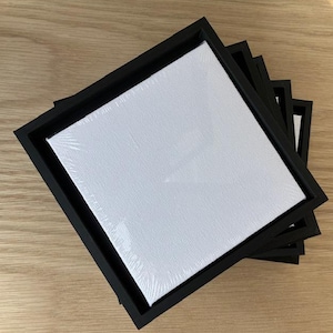 May include: A stack of square, white canvas panels in black wooden frames. The canvases are wrapped in clear plastic. The frames are black and have a beveled edge. The items are stacked on a light wood surface.