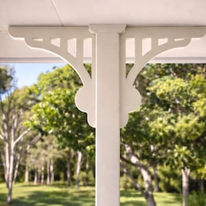 May include: A white porch post with decorative brackets. The brackets have a repeating pattern of cut-out shapes and rounded edges. The background shows a green lawn and trees, suggesting an outdoor setting.