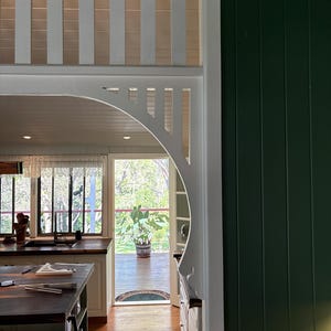 May include: Interior shot of a kitchen and outdoor deck. The kitchen has a dark wood island and worktops, with white cabinets and trim. The doorway has a decorative white arch and vertical slats. The outdoor deck has a potted plant.