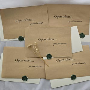 May include: Five rectangular kraft paper envelopes with black printed text reading Open when... followed by specific prompts including you need a laugh, you want to remember us, you miss me, you need inspiration, and you're stressed, with green wax seals.