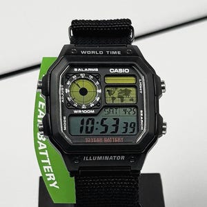 May include: Black Casio digital watch with a square face and black fabric band. The watch displays the time, date, and world time functions. The watch has the text "WORLD TIME", "CASIO", and "ILLUMINATOR" printed on the face.