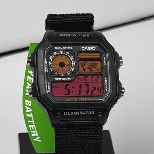 May include: Black Casio digital watch with a square face and a black fabric band. The watch displays the time in red digits and features multiple functions, including world time and alarms. The watch has the text "10 YEAR BATTERY" and "ILLUMINATOR".