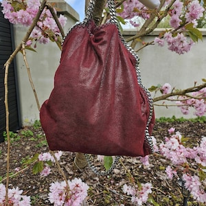 May include: A deep red shoulder bag with a silver chain strap. The bag is made of a textured material and is suspended from a tree branch with pink blossom. The bag has a drawstring closure.