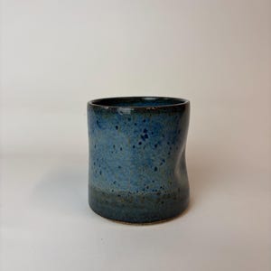 May include: A handmade ceramic tumbler with a speckled blue glaze. The cup has a slightly irregular shape and a dark blue rim. The base of the cup is a darker shade of blue. The tumbler is suitable for beverages.