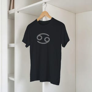 Może przedstawiać: Czarny t-shirt z szarym symbolem zodiaku Raka. Koszulka z kr&oacute;tkim rękawem wisi na drewnianym wieszaku. Tło to biała szafa.
