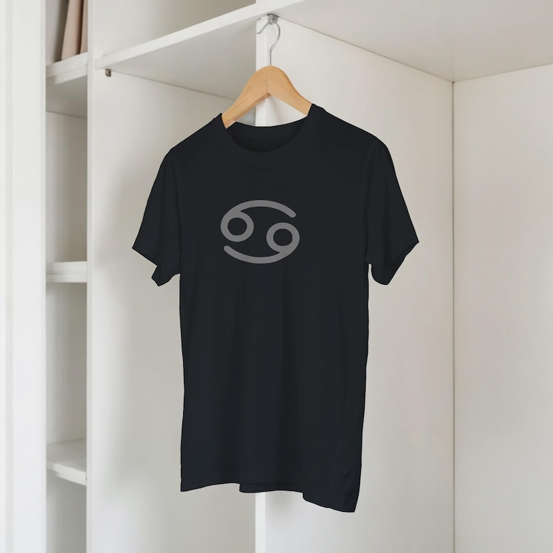 Może przedstawiać: Czarny t-shirt z szarym symbolem zodiaku Raka. Koszulka z kr&oacute;tkim rękawem wisi na drewnianym wieszaku. Tło to biała szafa.
