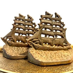 May include: Two bronze-colored ship bookends. Each bookend features a detailed sailing ship design with multiple masts and sails. The ships are mounted on a textured base, and the overall aesthetic is vintage.