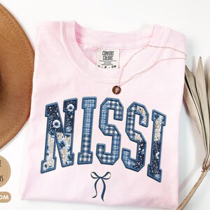 May include: A light pink t-shirt with the name NISSI embroidered in large block letters across the chest. The letters feature a mix of blue floral and blue gingham patterns. A small blue bow is embroidered below the name. A Comfort Colors tag is visible.