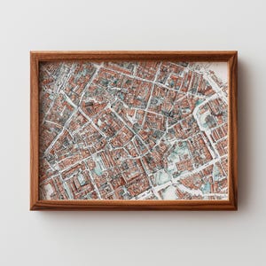 May include: A framed watercolor map print featuring a detailed aerial view of a city. The map is rendered in shades of red, gray, and blue, with a focus on the intricate network of streets and buildings. The print is encased in a brown wooden frame.