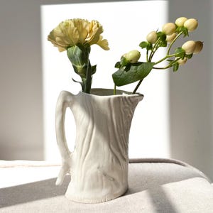 May include: A white ceramic pitcher with a textured, bark-like design, holding a yellow carnation and a sprig of green berries. The pitcher has a handle and a small spout. The arrangement is set against a white background.