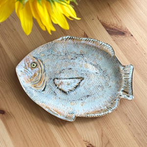 May include: A ceramic dish shaped like a fish with a light blue and white glazed finish. The surface features textured scales, a defined fin, and a prominent eye. The edges and details are highlighted with a rustic brown wash, creating a weathered look.