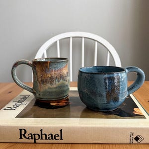 May include: Two ceramic mugs with a glossy finish. The left mug features a blend of blue, brown, and tan glaze with a tall, cylindrical shape. The right mug is a shorter, rounded bowl-like shape with a deep blue and dark brown mottled glaze.