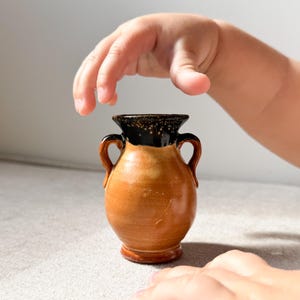 以下が含まれることがあります： 茶色のボディと黒い縁を持つ、手作りの小さなセラミック花瓶。花瓶には2つの小さな取っ手があり、丸みを帯びた形をしています。画像には、子供の手が花瓶に伸びている様子が写っています。