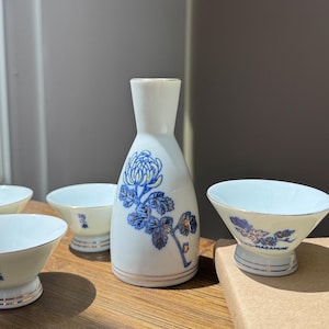May include: A white ceramic sake set featuring a bottle and four cups. The bottle and cups are decorated with blue floral designs and gold trim. The sake set is displayed on a wooden surface.