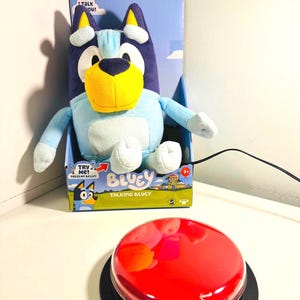 May include: A plush Bluey toy in its original box, featuring a light blue body, dark blue accents, and a yellow muzzle. The box reads "Talking Bluey" and "Try Me! Press my belly!" A large red button sits in the foreground.
