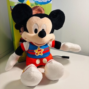 May include: A plush Mickey Mouse toy with large black ears, a red and blue outfit, and white gloves and shoes. The toy is in front of a box with the words "MICKEY MOUSE CLUBHOUSE" and "SINGING FUN" printed on it.