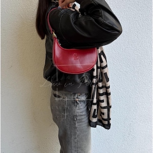 May include: A red leather crescent-shaped handbag with a gold-tone handle. The bag has a small embossed emblem and a curved design. The bag is held by a person wearing a black jacket and grey jeans.
