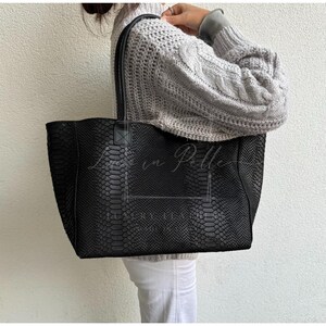 May include: Black tote bag with a snakeskin pattern. The bag has two handles and a rectangular shape. The text "Love in Pelle" is visible on the front of the bag. The bag is made of a textured material.