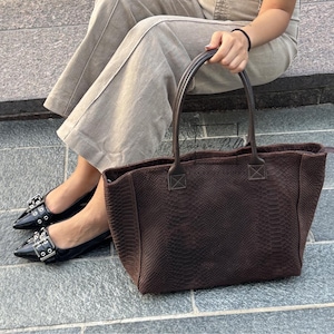 May include: A brown snakeskin-patterned tote bag with two handles. The bag is held by a person wearing black flats with silver buckles and light-coloured trousers. The bag appears to be of a medium size and is a dark brown colour.