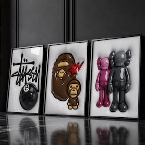 Peut inclure: Trois cadres muraux rectangulaires présentant des graphismes brillants en relief. Le premier montre un logo Stussy noir et une boule de billard numéro 8. Le deuxième affiche une tête de singe marron et un petit singe. Le troisième montre deux personnages KAWS.