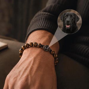 May include: A beaded bracelet featuring brown tiger eye stones and a central circular charm. The charm contains a small, circular projection lens that displays a clear, color photograph of a black Labrador retriever with its tongue out.