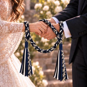 Puede incluir: Un cordón decorativo para ceremonia de unión con trenzas en azul marino, blanco y negro entrelazadas con delicados detalles de encaje floral. El cordón termina en dos borlas grandes y gruesas hechas de tiras de tela a juego en azul marino, blanco y negro.