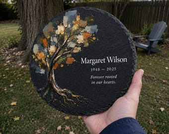 Puede incluir: Una placa conmemorativa redonda de pizarra negra con un árbol pintado de hojas doradas, marrones y grises. El texto dice Margaret Wilson, 1948 - 2025, y Forever rooted in our hearts. La placa tiene un borde texturizado y rugoso.