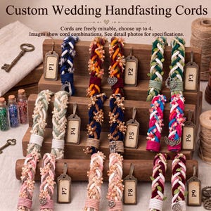 May include: Custom wedding handfasting cords displayed on a wooden stand. The cords are braided in various color combinations, including blue, green, pink, and red, featuring small charms and beads. Metal tags labeled P1 through P9 are attached.