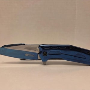 May include: A blue folding knife with a silver blade. The knife has a blue handle with a textured design and the text "MTech USA" is visible on the blade. The knife is open and ready for use.