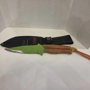 May include: A survival knife with a serrated blade and a green handle. The handle is wrapped in brown cord, and the knife comes with a black sheath. The knife has a total length of approximately 25 cm.