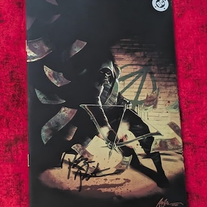 May include: A comic book cover featuring a hooded figure holding a bow and arrow, surrounded by floating paper currency. The artwork includes a DC logo in the top right corner, a brick wall background, and a black signature on the lower portion.